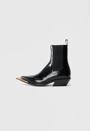 Bottine en cuir noir brillant avec des panneaux latéraux en élastique, bout pointu avec un embout en métal doré distinctif, et petit talon incliné sur fond blanc.