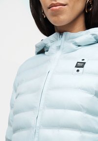 Κοντινό πλάνο ενός ατόμου που φοράει ανοιχτό μπλε μπουφάν με ραφές, κουκούλα και φερμουάρ, όπου φαίνεται η ετικέτα της μάρκας στο στήθος και χρυσά κρίκοι σκουλαρίκια.