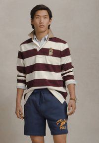 Polo cu mânecă lungă în dungi maronii și albe, cu guler, asociat cu pantaloni scurți bleu marin cu text galben, toate pe un fundal neutru.