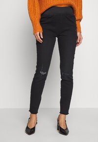 Jean skinny noir avec des détails déchirés au niveau des genoux, confectionné en denim. Porté avec un pull en tricot orange et des talons noirs, avec un ourlet effiloché.