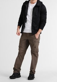 Zwarte ritsjack over een wit shirt, tan cargobroek met ritsen en logo, zwarte wandelschoenen. Glad materiaal met een casual fit.