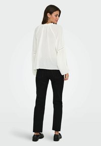 Žena oblečená v bílé průhledné blůze s dlouhými rukávy a tečkovanou texturou, s černými džínami, stojící zády k fotoaparátu.