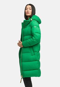 Femeie cu păr drept negru, îmbrăcată într-o haină lungă, căptușită, de culoare verde strălucitor, cu glugă, stând în poziție de trei sferturi pe un fundal alb.