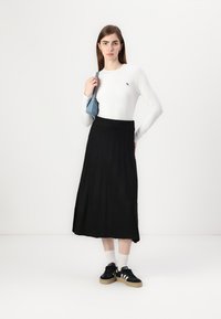 Hvit langermet topp, svart plissert skjørt og svarte sneakers med hvite detaljer. Modellen holder en liten blå veske. Enkel hvit bakgrunn.