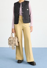 Chaleco acolchado negro sobre una camisa de manga larga rosa claro, combinado con pantalones beige de cintura alta y zapatos negros gruesos. Bolso beige con detalles.