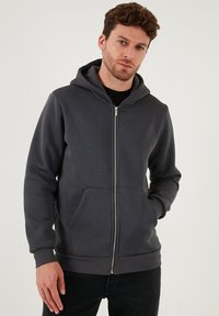 Sivi dukserica na patent, s kapuljačom s vezicom, prednjim džepovima, rebrastim manžetama i glatkom teksturom. Nosena preko crne majice.