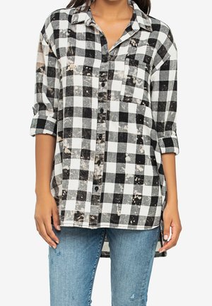 Femme portant une chemise à manches longues à carreaux noir et blanc avec les manches retroussées et des taches décolorées, associée à un jean bleu clair déchiré.