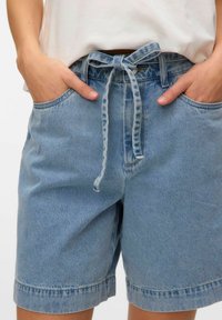 Šviesiai mėlyni džinsiniai šortai su raišteliu juosmens srityje, dviem šoninėms kišenėms ir pasukta apdaila. Audinys turi lygų paviršių.