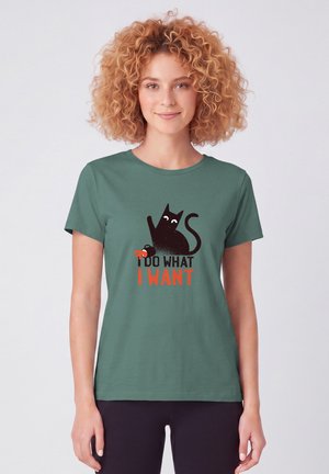 Jeune femme aux cheveux roux bouclés portant un t-shirt vert avec un chat noir et le texte "I DO WHAT I WANT."