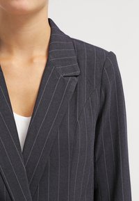 Blazer en velours côtelé marine, avec un col à revers, coupe ajustée et tissu texturé. Caractéristiques des revers nets et des détails de couture subtils.
