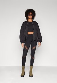 Musta bomber-takki, jossa on röyhelövetoketju, musta crop-top, mustat legginsit, joissa on pitsileikkaukset, ja oliivinvihreät korkokengät neliön muotoisella kärjellä.