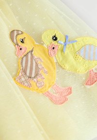 Gult tyg med två broderade ankungar med rosa och blå accenter. Den ena har randig rygg och en ljusblå rosett.