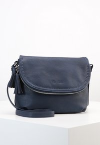 Marineblå læder crossover taske med et fold-over låg, sølv lynlås, frynse dekorationer og justerbar skulderrem præsenteret på en hvid overflade.