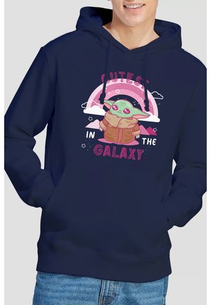 Person wearing navy hoodie with cartoon green alien in brown coat, pink rainbow, stars, and text "Cutest in the Galaxy."