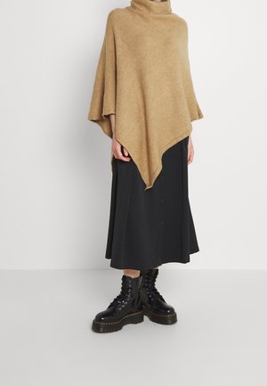 Personne portant un poncho marron, une jupe midi noire avec des boutons et des bottes plateforme noires à lacets, debout contre un mur blanc uni.