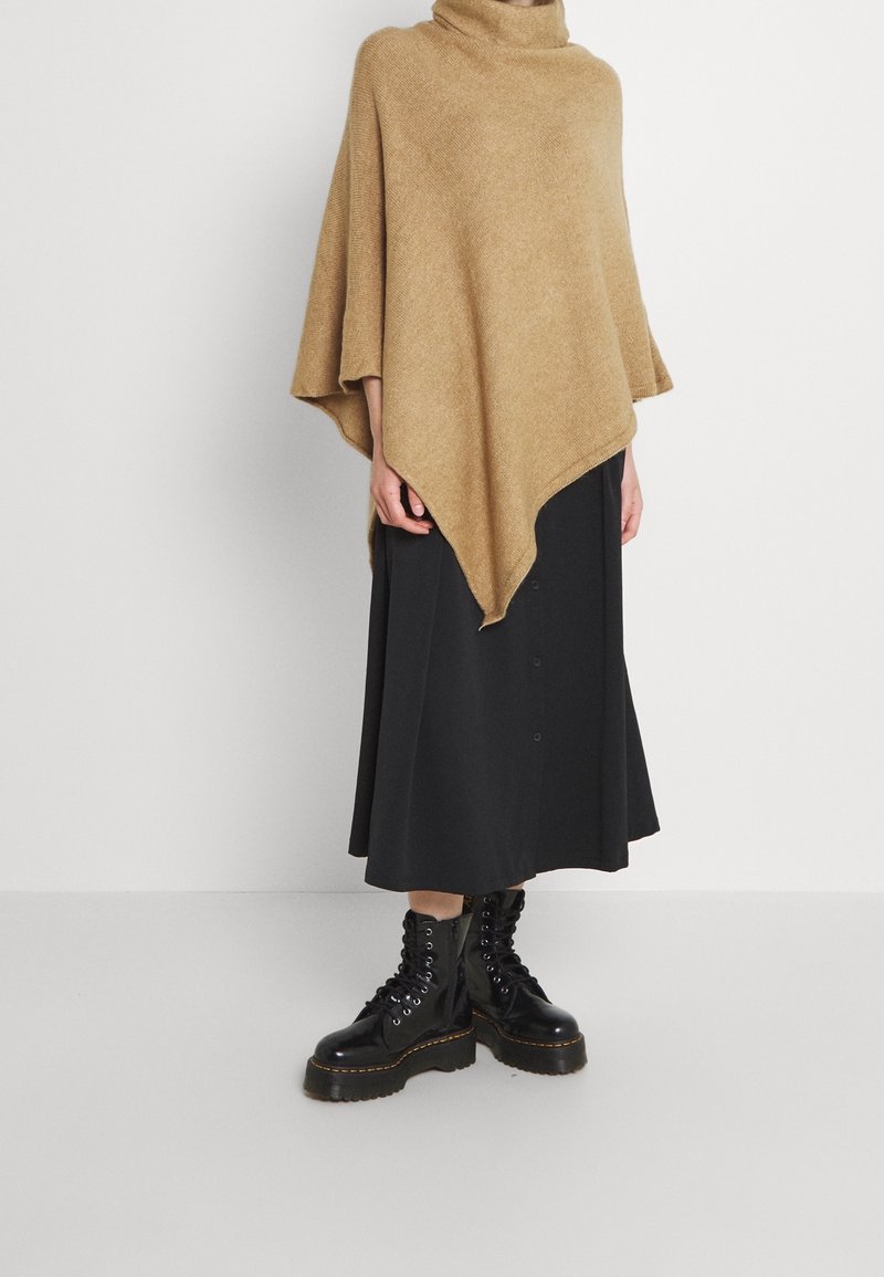 Personne portant un poncho marron, une jupe midi noire avec des boutons et des bottes plateforme noires à lacets, debout contre un mur blanc uni.