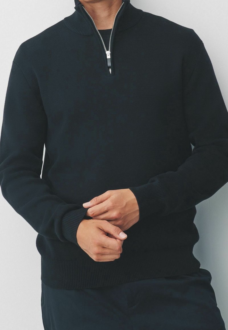 Pull en maille noir avec un col montant à fermeture éclair, des poignets côtelés et un ourlet droit. Le tissu a une texture lisse.