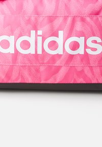Rosa Adidas-merket veske med teksturert stoff og blomstermønster, stor hvit logo og en kontrastfylt svart bunn.