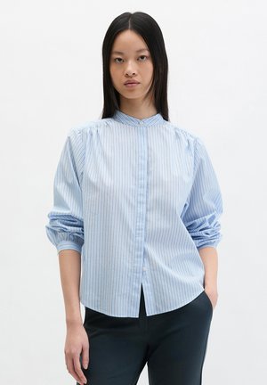 Młoda kobieta o długich czarnych włosach, ubrana w jasnoniebieską koszulę w paski z zebranymi rękawami i czarne spodnie na białym tle.