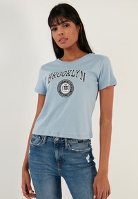Gaiši zils apgriezts t-krekls ar uzrakstu "BROOKLYN" un apļa grafikas elementu. Ar īsām piedurknēm un apaļu kakla izgriezumu. Apvienots ar ziliem džinsiem.