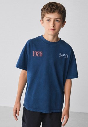 Junge mit lockigem Haar, der ein übergroßes navyblaues T-Shirt mit den Logos "1988" und "BAKER by Ted Baker" trägt, steht vor einem schlichten hellen Hintergrund.