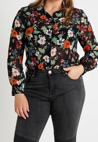 Femme portant un haut floral transparent noir avec des fleurs rouges, blanches et vertes, associé à un jean taille haute gris foncé, la main dans la poche.