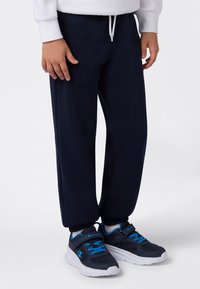 Barn som har på seg marineblå joggebukser og marineblå joggesko med blå lisser og hvite såler, står på en hvit bakgrunn.