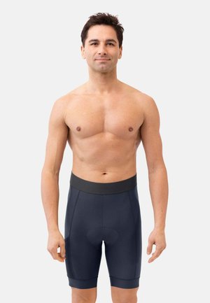 Jeune homme debout torse nu portant un short de cyclisme noir sur un fond clair uni.