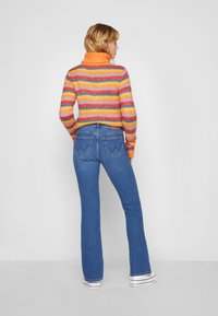 Fargerik stripete høyhalset genser med oransje, gul og lilla nyanser, paret med tettsittende blå utsvingte jeans med baklommer med sømdetaljer.