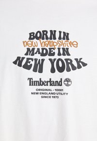 Hvid bomulds T-shirt med sort og orange grafisk tekst: "Født i New Hampshire, lavet i New York," med Timberland-logo og detaljer nedenfor.