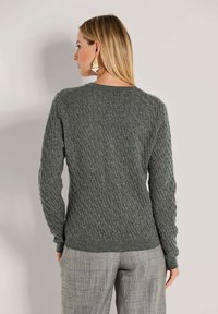 Frau mit blonden Haaren, die einen grauen Kabelstrickpullover und graue Karohosen trägt, mit dem Rücken zu einer einfarbigen, hellen Hintergrund.
