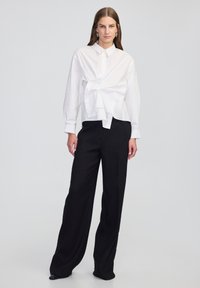 White long-sleeve shirt with a front tie detail, paired with black high-waisted, wide-leg trousers, presented against a neutral background.
