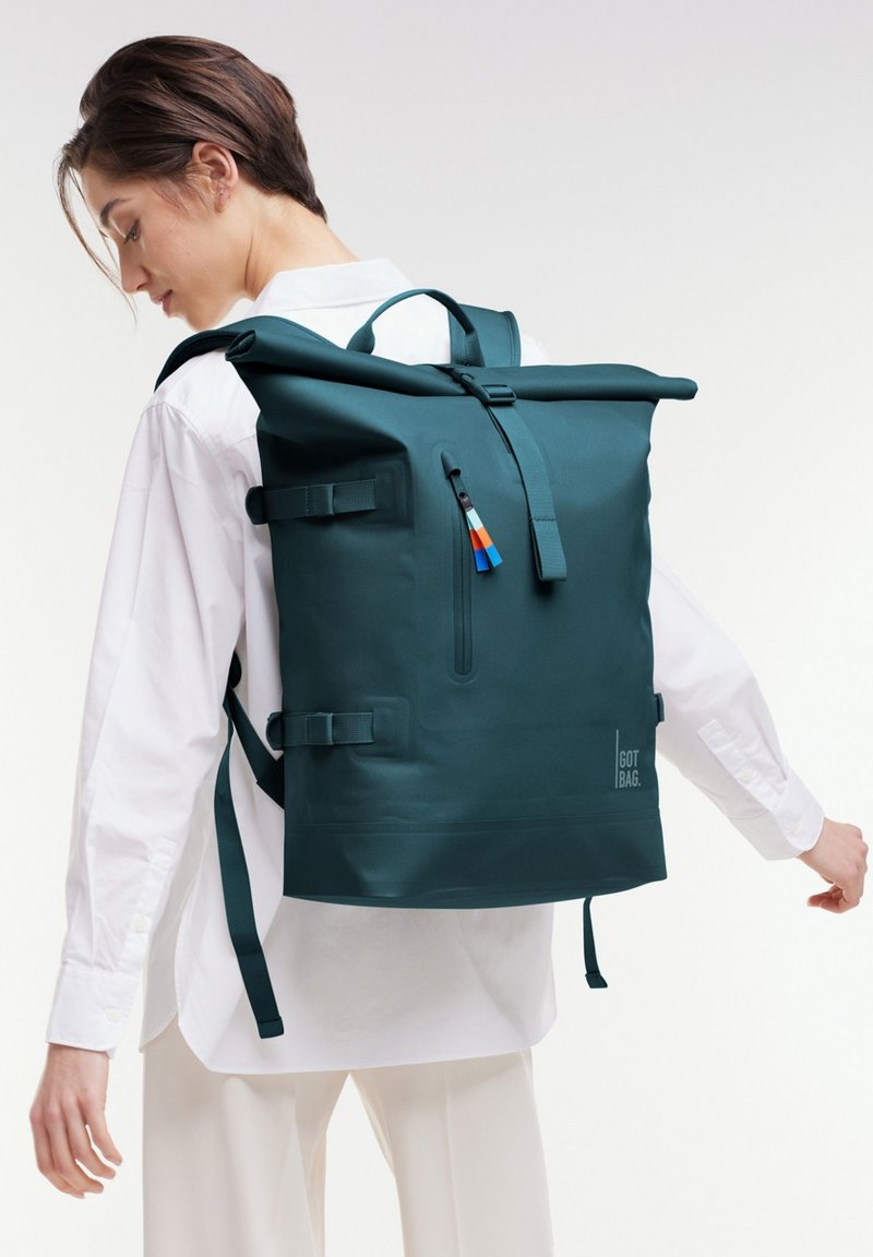 Teal roll-top reppu, joka on valmistettu kestävästä materiaalista. Varusteena on säädettävät hihnat, etutasku vetoketjulla ja värikkäitä varusteita.