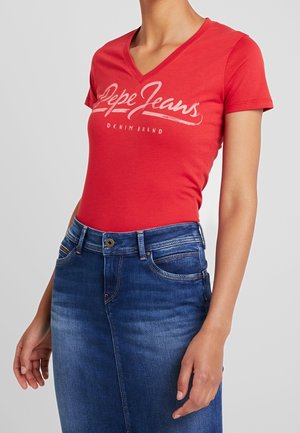 Kvinde iført en rød V-hals Pepe Jeans T-shirt, stukket ned i et højtaljet blåt denimskørt med forlommer og bælteløkker.