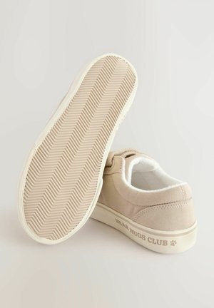 Scarpe di tela beige con una suola in gomma bianca testurizzata, dotate di rivestimento morbido e con la scritta "BEAR HUGS CLUB" stampata sul lato.