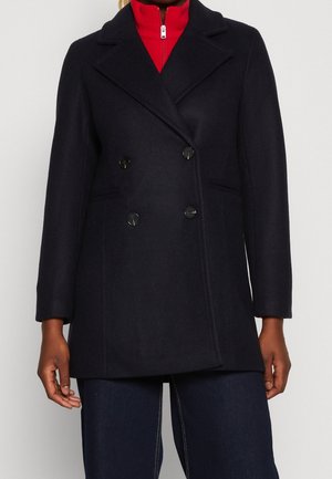 Personne portant un manteau noir à double boutonnage sur un haut rouge zippé et un pantalon bleu foncé, debout devant un fond clair uni.