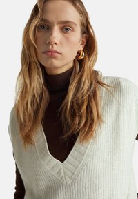 Cremefarbener gestrickter Vest mit weitem V-Ausschnitt und gerippten Abschlüssen, getragen über einem dunkelbraunen Rollkragenpullover. Lange, gewellte Haare und kreisförmige goldene Ohrringe sind zu sehen.