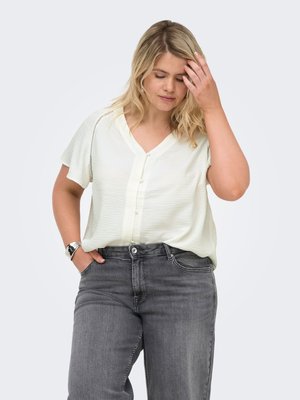 Femme blonde portant un chemisier blanc manches courtes et un jean gris, une main dans la poche et l'autre touchant ses cheveux.
