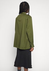 Camicia a maniche lunghe di colore verde oliva, in tessuto strutturato, con taglio dritto e spalla posteriore. Abbinata a una gonna nera con orlo sfrangiato.