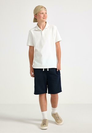 Dreng iført beige kasket bagud, hvid poloshirt, navy shorts, hvide sokker og beige sneakers, stående og kigger til siden.