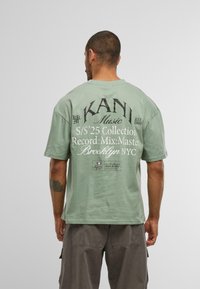 Meldrzaļš kokvilnas krekls ar īsām piedurknēm; aizmugurē ir lieli melni burti un grafikas, tostarp "KANI Music" un kolekcijas detaļas.