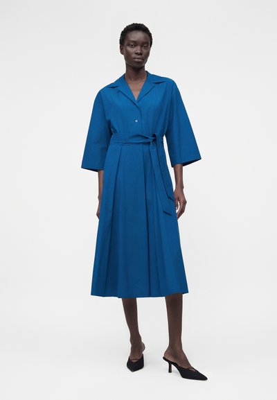 Γυναίκα με μπλε midi φόρεμα με φαρδιά μανίκια, γιακά, μπροστινά κουμπιά, ζώνη δεμένη στη μέση και μαύρα μυτερά τακούνια, όρθια μπροστά σε λευκό φόντο.