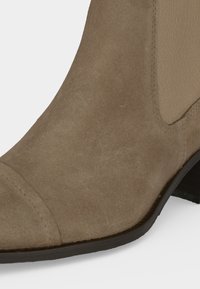 Gros plan sur une botte Chelsea en daim beige montrant une pointe cousue, un panneau latéral élastique et un petit talon noir.