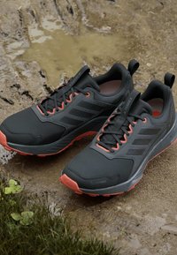 Paar schwarze und orangefarbene Trailrunning-Schuhe mit griffigen Sohlen, die auf schlammigem Boden in der Nähe von Gras stehen.