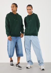 Mørkegrønne hettegensere med kengurulommer, kombinert med lyseblå vide jeans. Sneakers har kontrasterende farger og stiler.