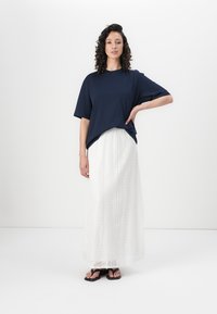 T-shirt navy oversize; gonna maxi bianca in pizzo con texture a fantasia; infradito nere. La modella sta in piedi con una mano sui fianchi.