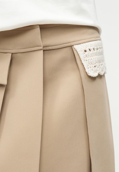 Beige skreddersydde bukser med høyt liv, med en hvit heklet lomme på høyre side.