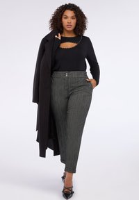 Žena s kudrnatými vlasy, oblečená v černém topu se cvočky, šedých kockovaných kalhotách s vysokým pasem, černém kabátu přehozeném přes rameno a černých střevících na špičku, kráčející vpřed.