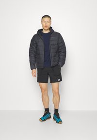 Giacca a vento nera con cappuccio, chiusura con zip e polsini a coste. T-shirt blu navy, pantaloni corti neri, abbinati a scarpe da ginnastica blu e nere.