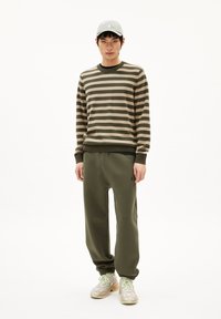 Gestreepte trui in olijfgroen en beige, gecombineerd met olijfgroene joggers. De model draagt een grijze pet en lichtgekleurde sneakers met pastelaccenten.